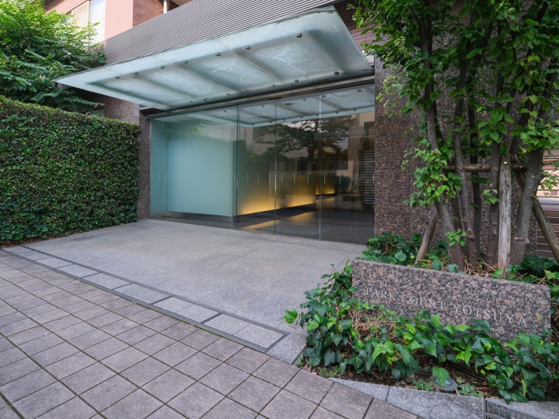 ガラス張りのエントランスには庇が設けられ、植栽の彩りが建物の表情を豊かに演出しています。上品な佇まいが印象的です。